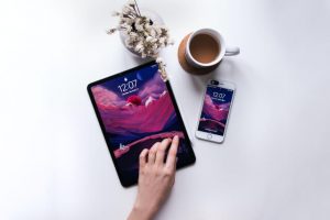 An ipad and iphone laying on a table with a cup of coffee and vase of flowers