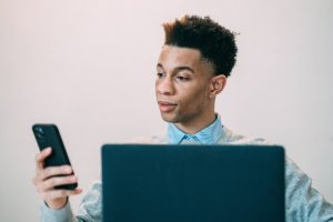 Man sitting behind open laptop, looking a smartphone in his hand