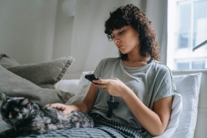 Woman sitting in bed petting a cat while looking at social media on her smartphone