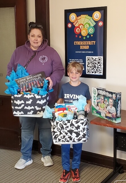 Woman and boy each holding a basket filled with games, snacks, and other prizes. The poster on the wall is advertising the Cybersecurity Bingo contest.