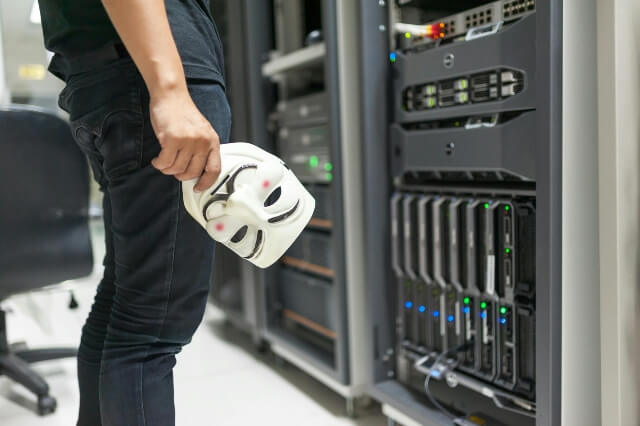 Man (view from waist down) standing in front of a server, holding an "anonymous" mask in his hand.