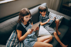 Two young kids sitting on the couch, playing on tablet devices