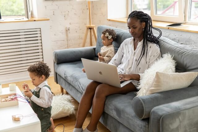 A remote worker sitting on her couch using a laptop. A small child is sitting on the couch with her and second child is playing with toys on the coffee table.