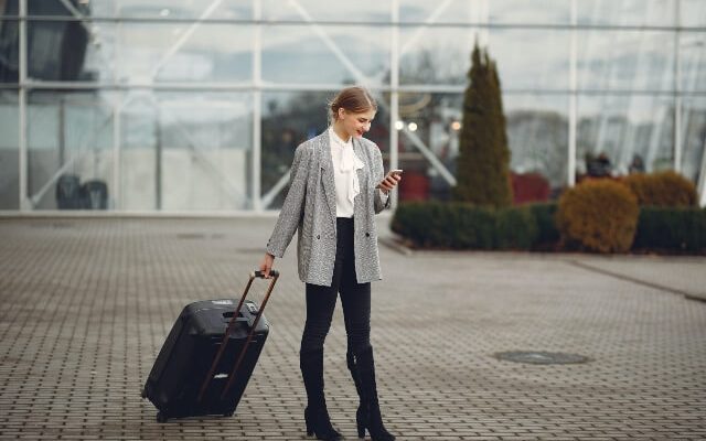 Woman standing at airport with suitcase, checking smartphone