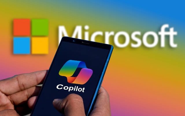 Hands holding a smartphone with the Microsoft Copilot logo. In the background is the Microsoft logo.