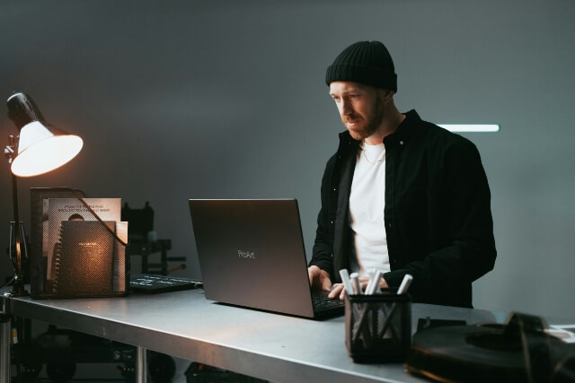 Man in dark clothes and a black beanie at a desk working on a laptop