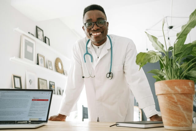 Doctor with a white coat and stethoscope standing behind a desk. On the desk are a laptop computer and a potted plant.