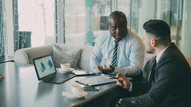 Two men in business attire sitting in front of a laptop computer, reviewing data.