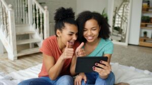 Two teenage girls smiling and giving a thumbs up to someone on a tablet