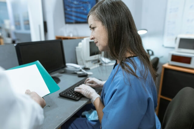 Nurse completing documentation on a laptop. She is looking at patient files being held by a doctor besider her. Her face shows concern.