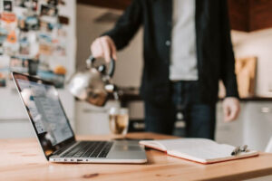 Man in his kitchen pouring hot water from a tea kettle. A work laptop and open notebook are on the table in the foreground.
