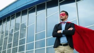 Man in a business suit with a red cape and red mask standing with crossed arms in front of an office building.